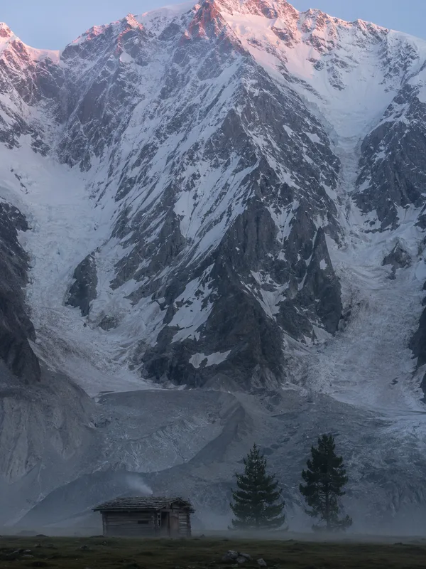 The Raikot face of Nanga Parbat filling the sky above the meadow, a wooden hut small on the grass below.