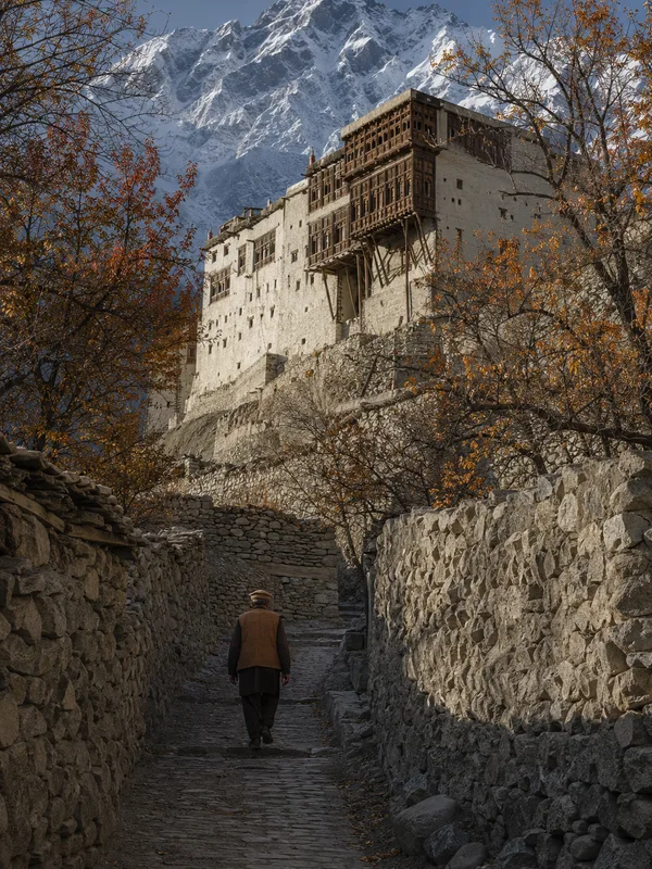 A man in a Hunza cap walking up a cobbled lane in old Karimabad towards Baltit Fort, Ultar Sar rising behind.