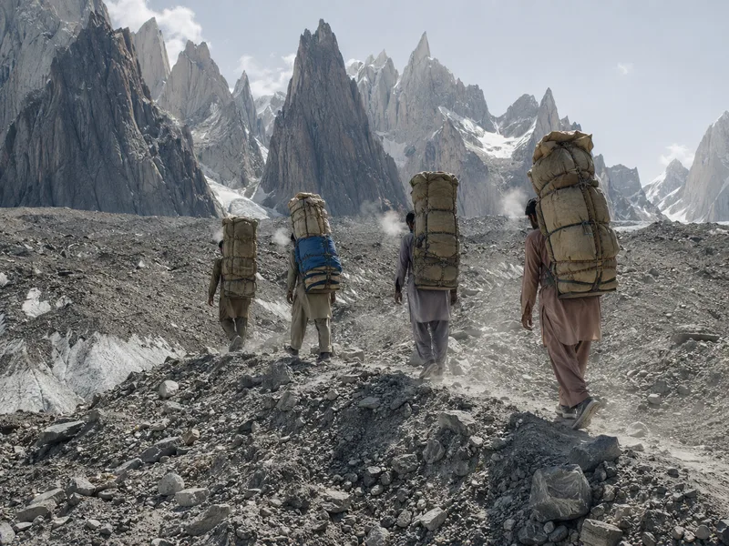 Balti porters carrying roped loads in single file across the moraine of the Baltoro glacier, granite spires behind.