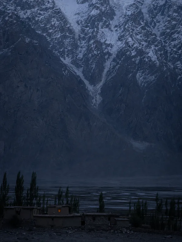A wall of Karakoram rock and snow towering over low Balti houses and poplars on the floor of the Skardu valley at dusk, one window lit.