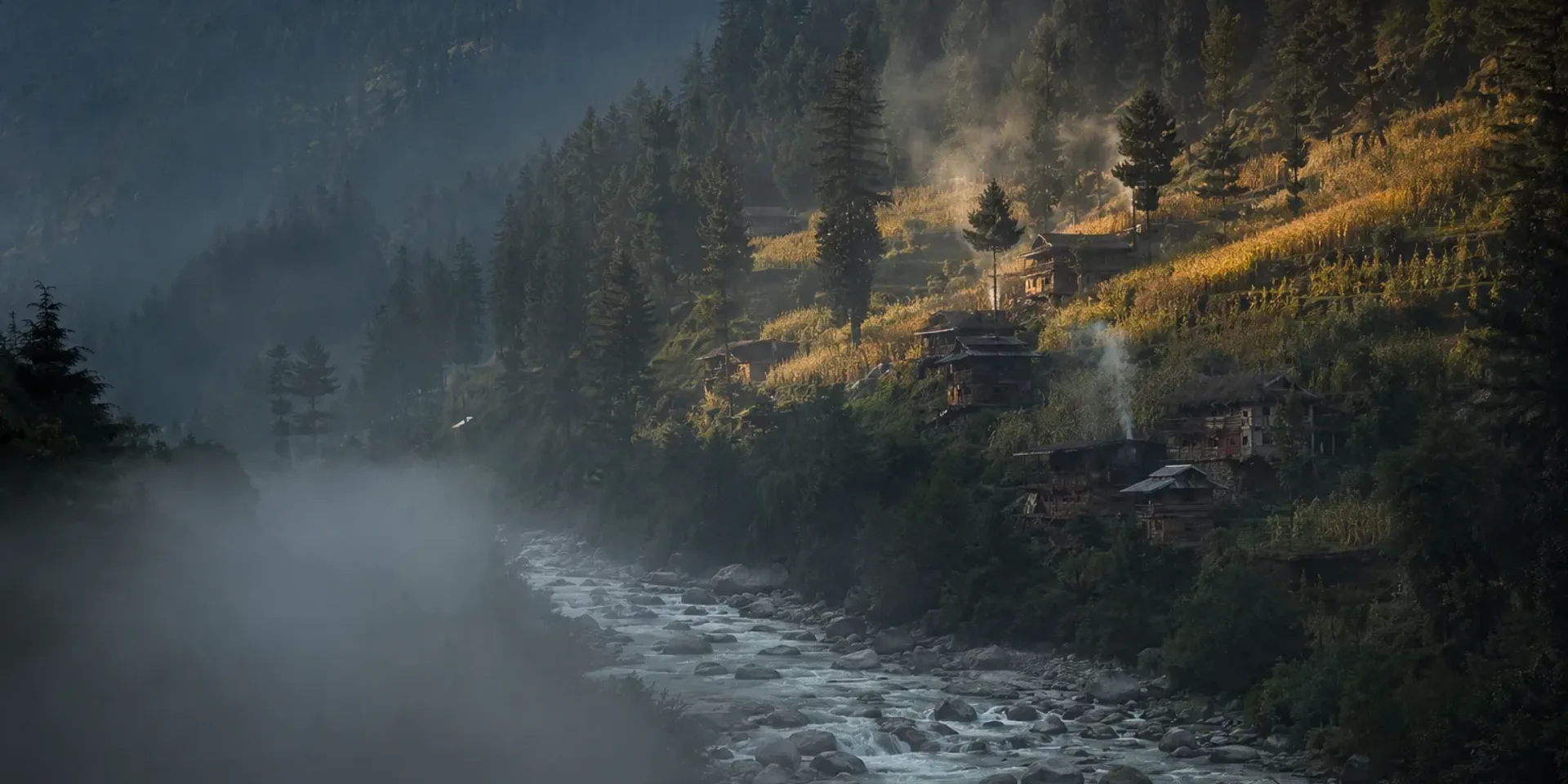Mist rising off the Swat river at dawn below wooden Kohistani houses and terraced fields on the forested valley side.