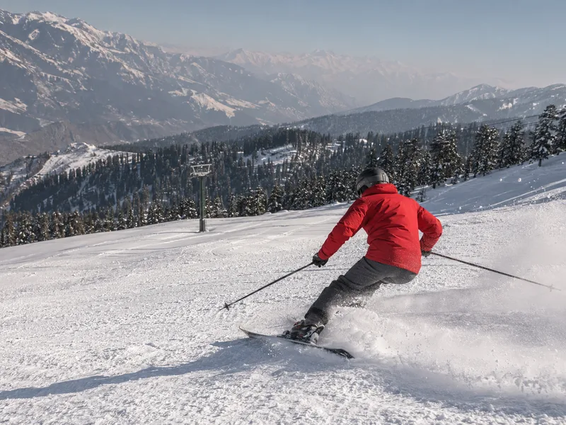 A skier in a red jacket carving through deep fresh powder at Malam Jabba, deodar forest below and peaks on the horizon.