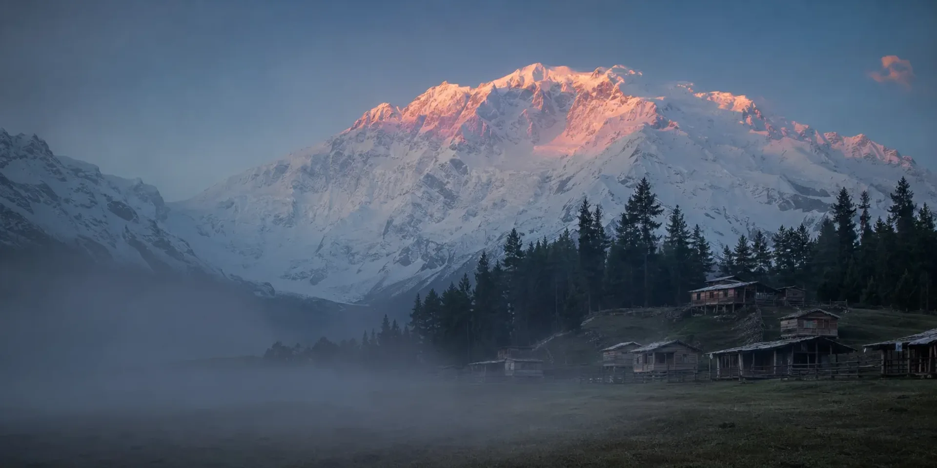 The meadow at Fairy Meadows at first light, wooden huts on the grass and the Raikot face of Nanga Parbat filling the sky behind.
