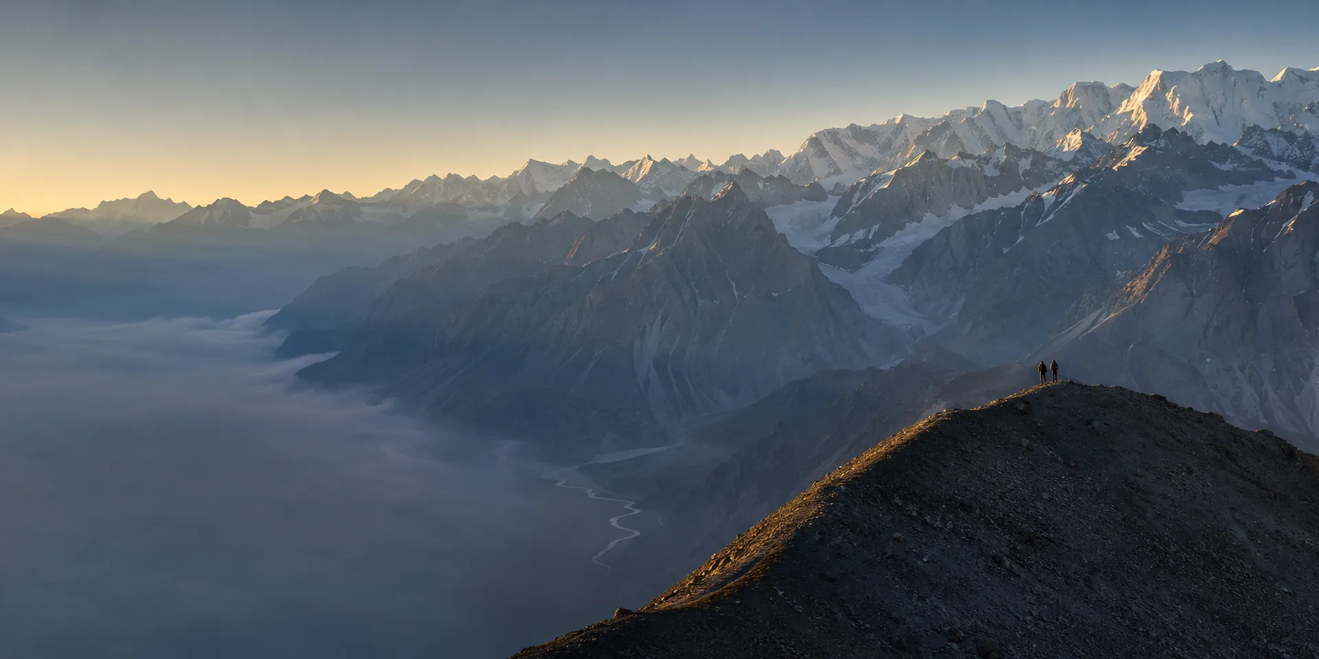 Two trekkers silhouetted on a high ridge above the Indus at sunrise, the Karakoram receding behind them in layers of snow peaks.