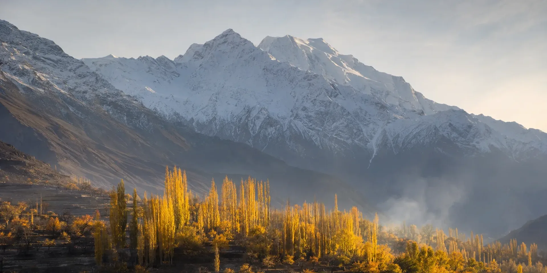 The Hunza valley in peak autumn colour, golden poplars and terraces below a snow wall in late-afternoon light.