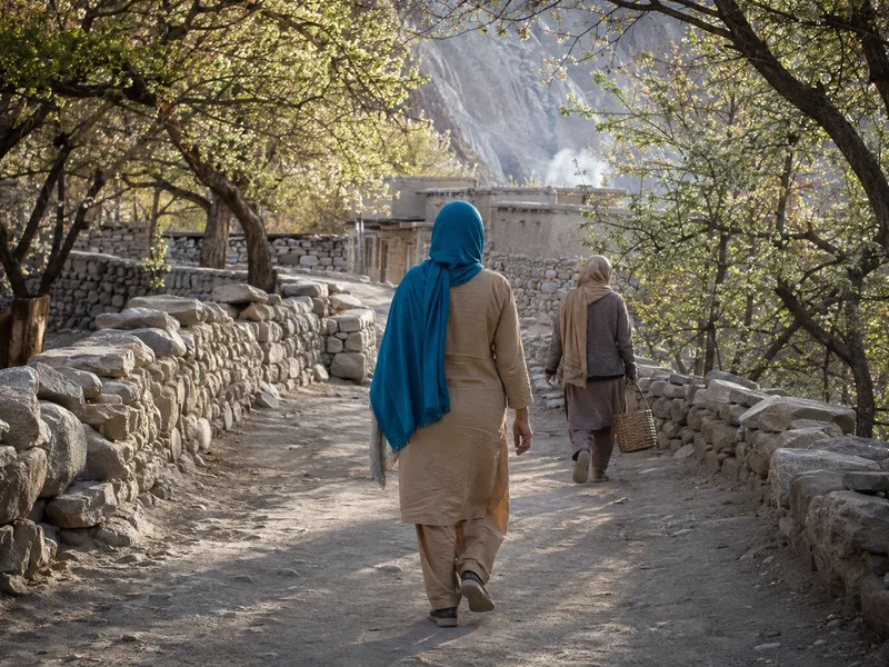 A woman traveller in a shalwar kameez and scarf walking a village lane in Hunza behind a local woman carrying a basket.
