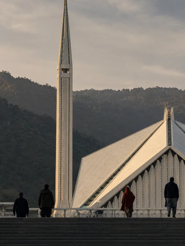 Faisal Mosque in Islamabad seen from below, its folded white roof and minaret against the wooded Margalla Hills.