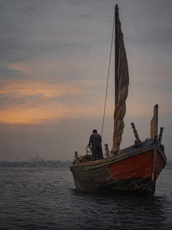 A wooden fishing boat with a tall mast on the Sindhi coast at dusk, the Karachi shoreline behind under a warm hazy sky.