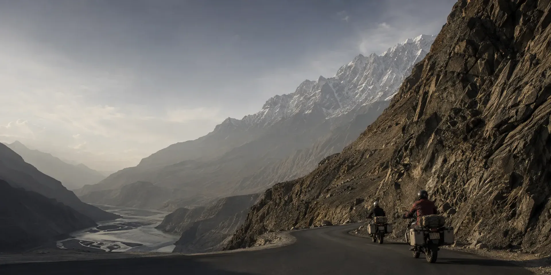 Two riders on loaded motorcycles on the Karakoram Highway where it curves along a cliff high above the Hunza river.