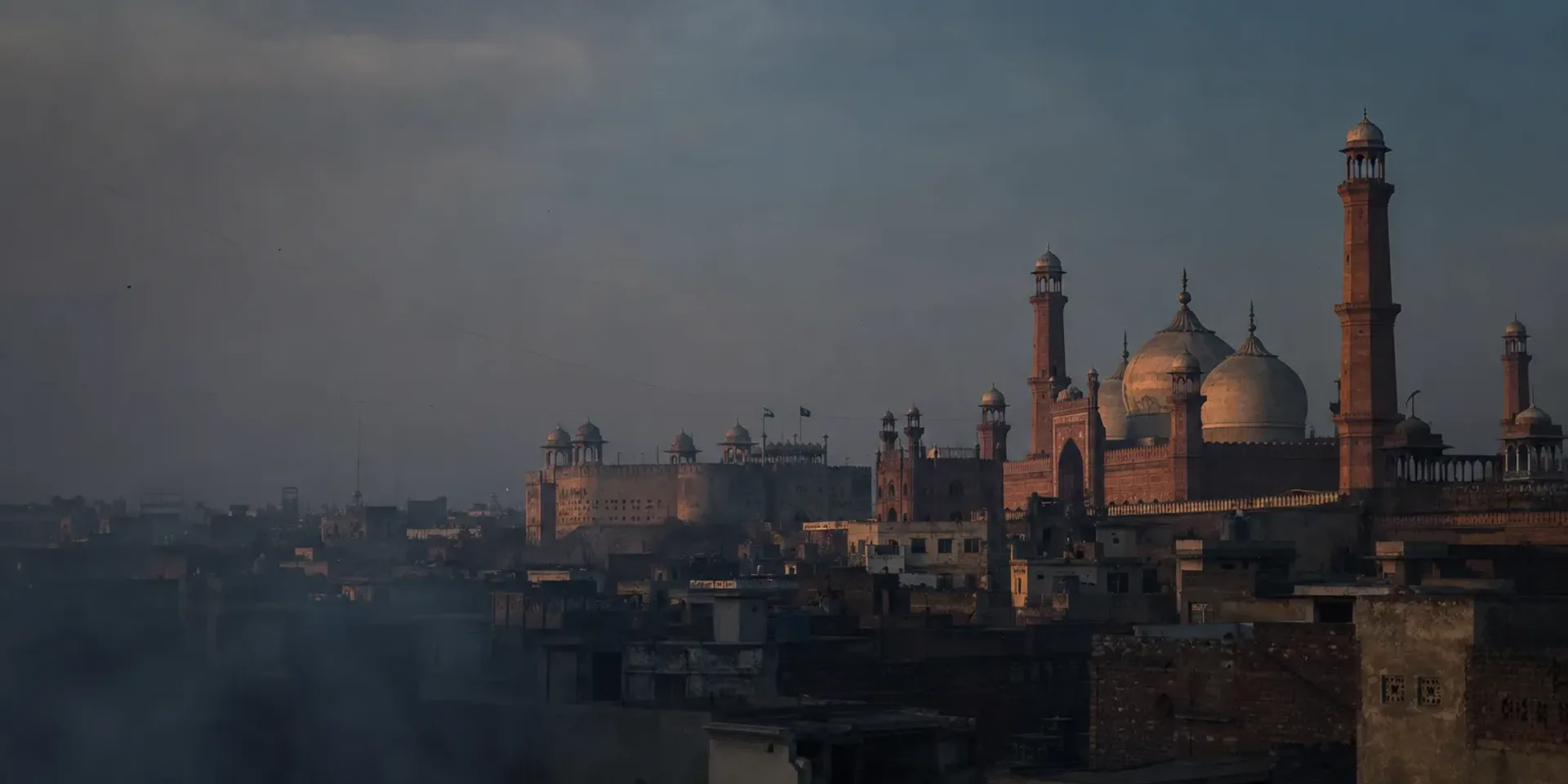 The Walled City of Lahore at dusk, domes and minarets above layered flat rooftops in a haze of woodsmoke.