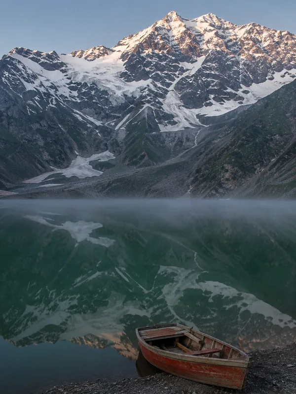 Lake Saif-ul-Malook at dawn, still water reflecting Malika Parbat with a wooden boat drawn up on the shingle.